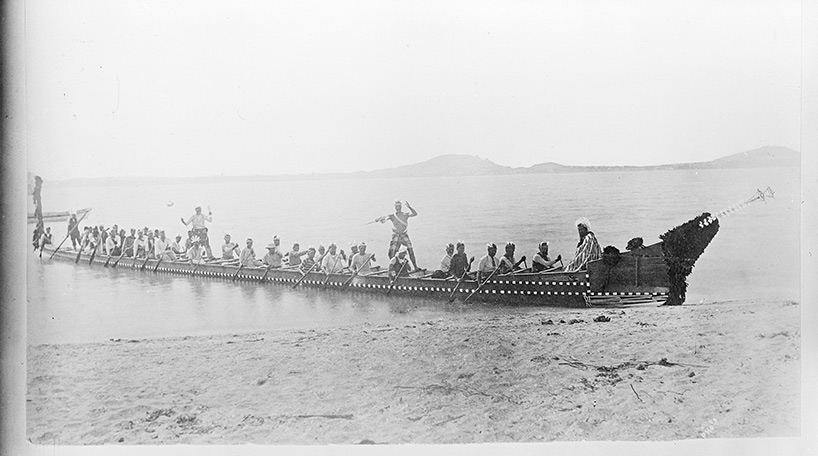 A black and white photo of people in a canoe on the shore. This is part of the Auckland Council Libraries heritage collections.