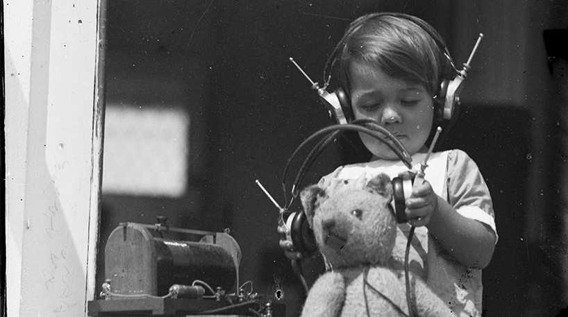 A black and white photo of a boy with headphones putting on another pair of headphones on a teddy bear. This is part of the Auckland Council Libraries heritage collections.