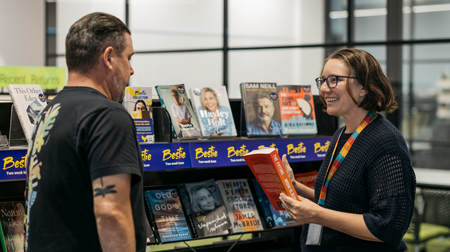 Smiling librarian holding a book talking to a library customer in front of a bookshelf