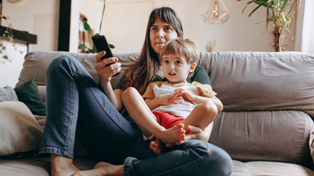 An adult sits on a couch with a child sitting on their lap. The adult is holding a television remote.