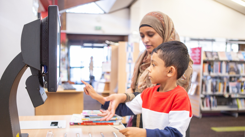 Child and parent borrowing books using a self-checkout machine at a library
