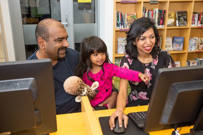 Image of a family of three using a computer at a public library. Their small child points at the screen with excitement.