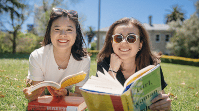 Photo of two young women reading books on the grass in the sun.