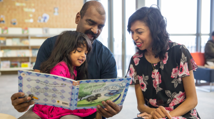 Two parents reading a picture book with a yound child in a library.