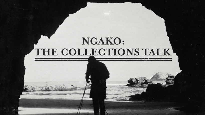 A black and white photo of a photographer in the mid-ground setting up a tripod at the mouth of a cave. The background is somewhat rocky with the ocean behind.