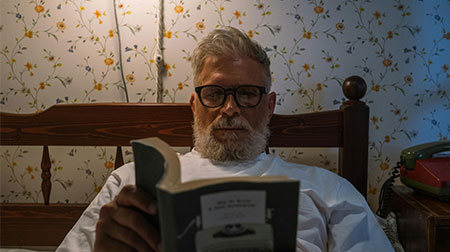 A man reads a book in bed by lamp-light.