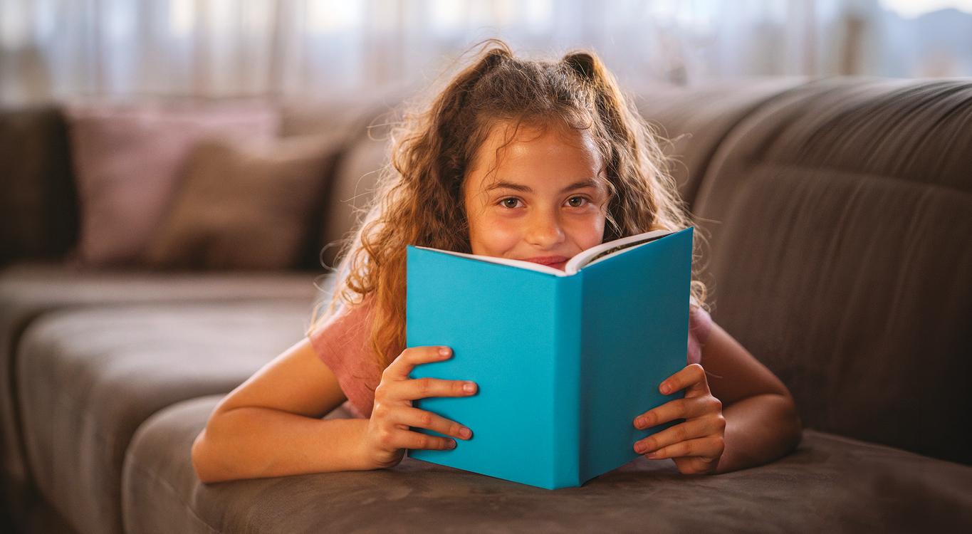 A young girl lying on the sofa and smiling over the top of a book cover.