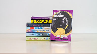 A stack of fiction books with one front-facing book cover titled They Dream in Gold