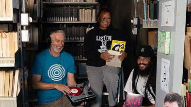 Brent, Nia and Julian holding vinyls amongst library basement shelves