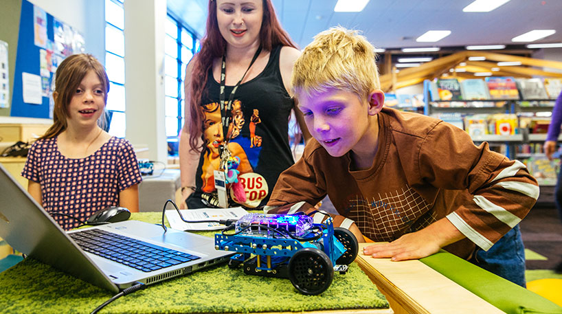 A girl watching a boy lean over a robotic car in a library.